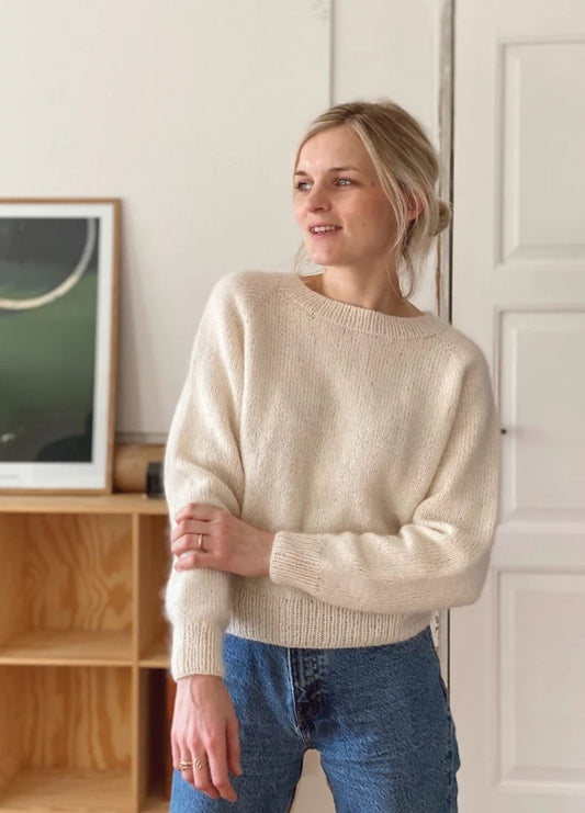 Woman in a cozy cream sweater and jeans stands indoors, smiling to the side. A framed picture and wooden shelf are visible in the background.
