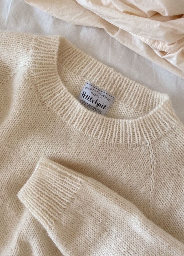 Cozy cream sweater with ribbed cuffs and neckline, labeled "PetiteKnit." It rests on a soft, light-coloured fabric, conveying warmth and comfort.