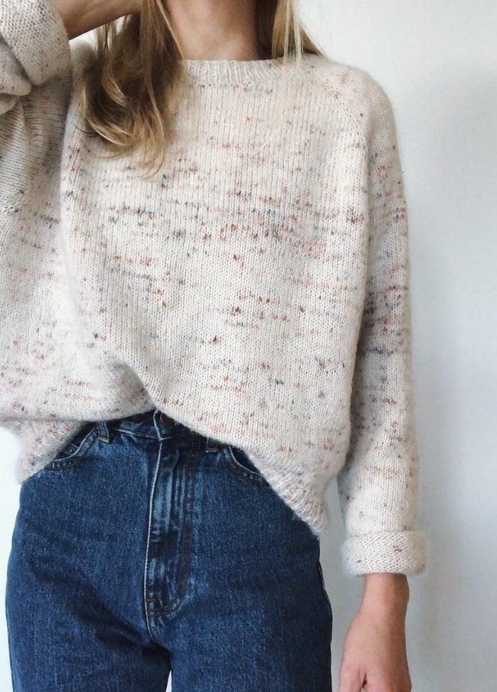 Woman in a cozy cream speckled sweater and high-waisted jeans stands against a plain background. The style is casual and relaxed.