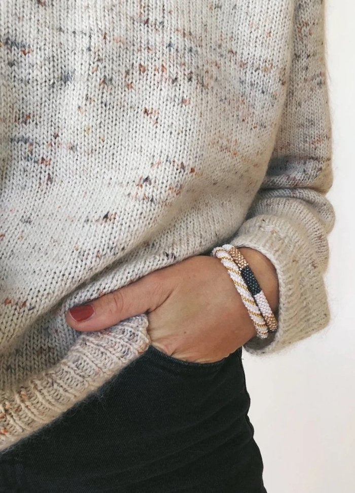 Close-up of a person wearing a speckled cream sweater, hand in a pocket, featuring beaded bracelets and dark jeans. The tone is cozy and casual.