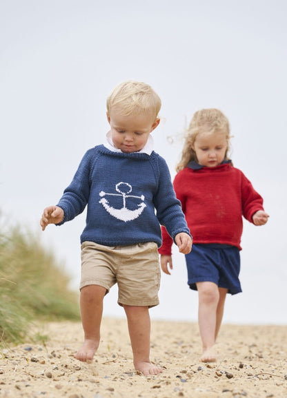 Two young children walk barefoot on a sandy path. The boy, wearing a blue sweater with an anchor, leads a girl in a red sweater. The mood is playful.