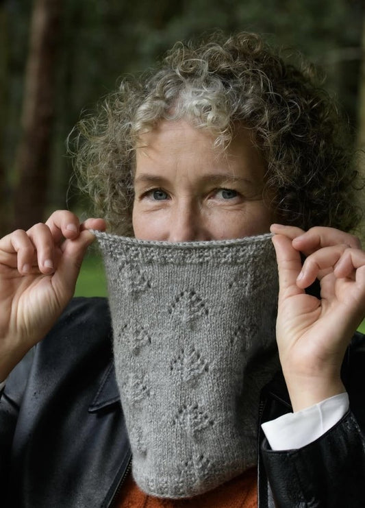 A person with curly hair holds a gray knitted scarf over their face, revealing only the eyes. The background is a blurred forest. The mood is cozy and playful.
