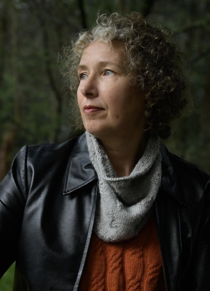 A woman with curly hair gazes thoughtfully to the side in a forest setting. She wears a black leather jacket, gray scarf, and orange sweater, conveying a contemplative mood.