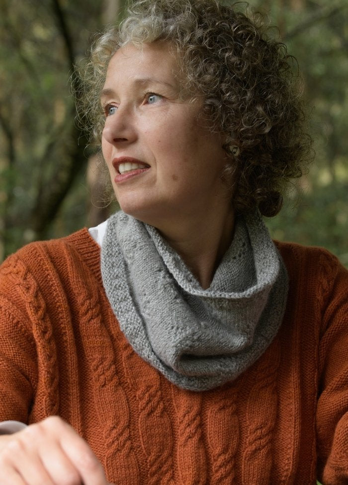 A woman with curly hair gazes to the side, wearing an orange sweater and a gray scarf. She appears thoughtful, set against a blurred forest backdrop.