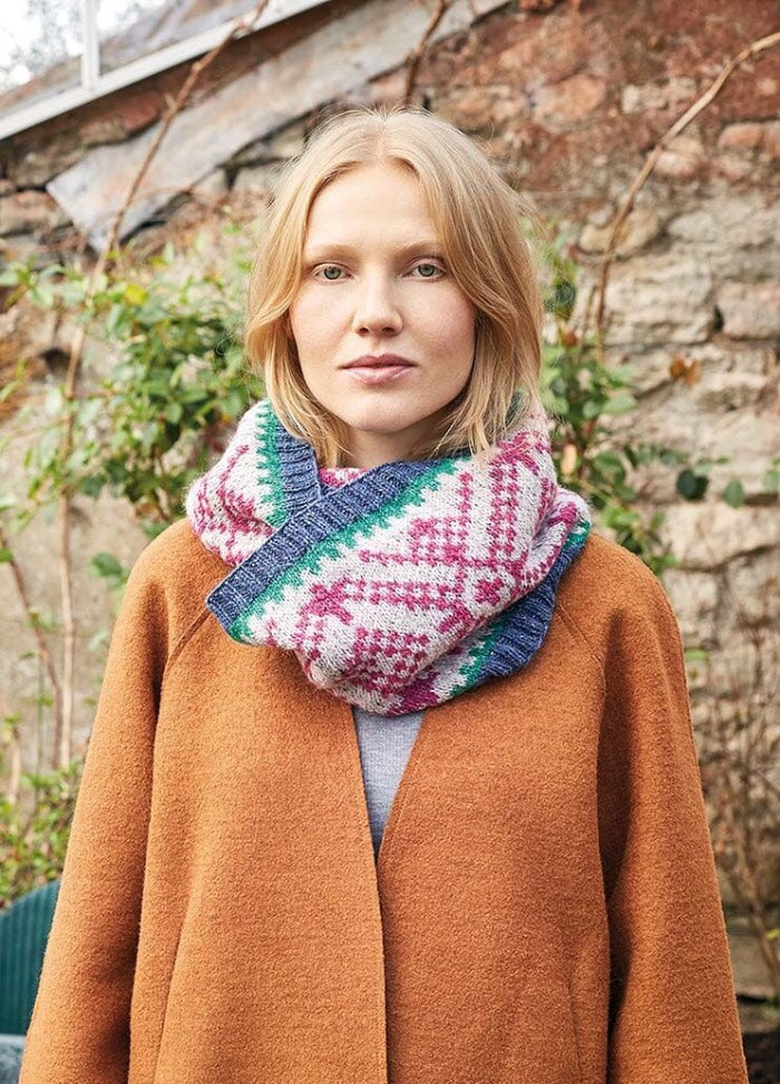 Woman in a rust-colored coat and multicolored knit scarf stands outdoors against a stone wall and greenery, conveying a serene autumn vibe.