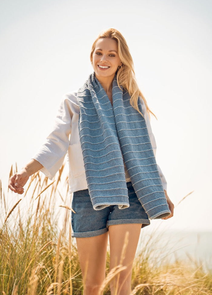 A person stands smiling in a sunny field of tall grass, wearing a blue striped scarf, white long-sleeve shirt, and denim shorts, creating a relaxed, cheerful vibe.