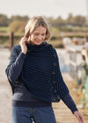 A woman smiles gently, wearing a textured navy blue poncho with buttons, standing on a wooden dock. She appears relaxed, with a blurred outdoorsy background.