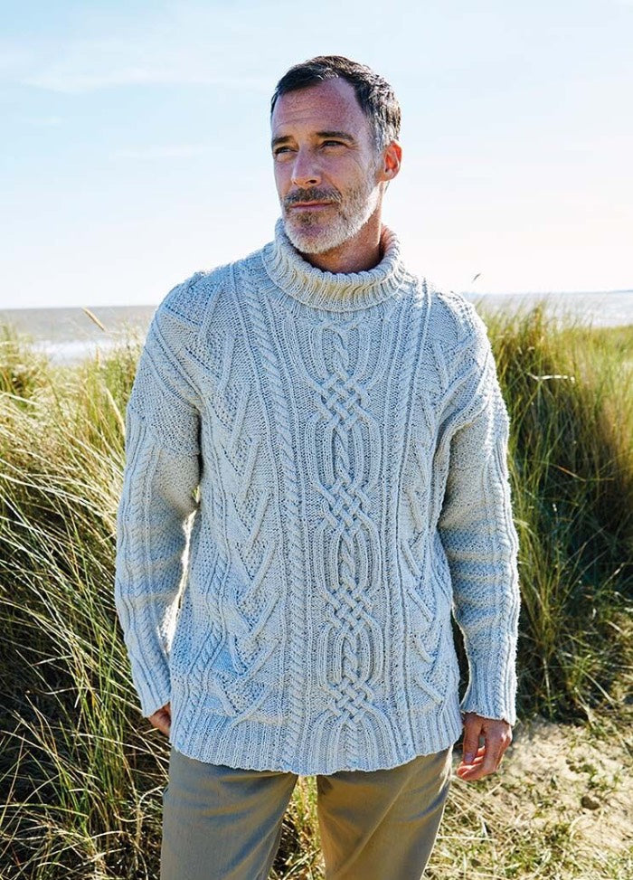 Man in a thick, cable-knit sweater stands in a grassy coastal landscape. He gazes into the distance, conveying a thoughtful, serene mood.