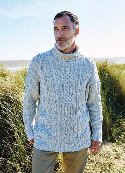 Man in a thick, cable-knit sweater stands in a grassy coastal landscape. He gazes into the distance, conveying a thoughtful, serene mood.