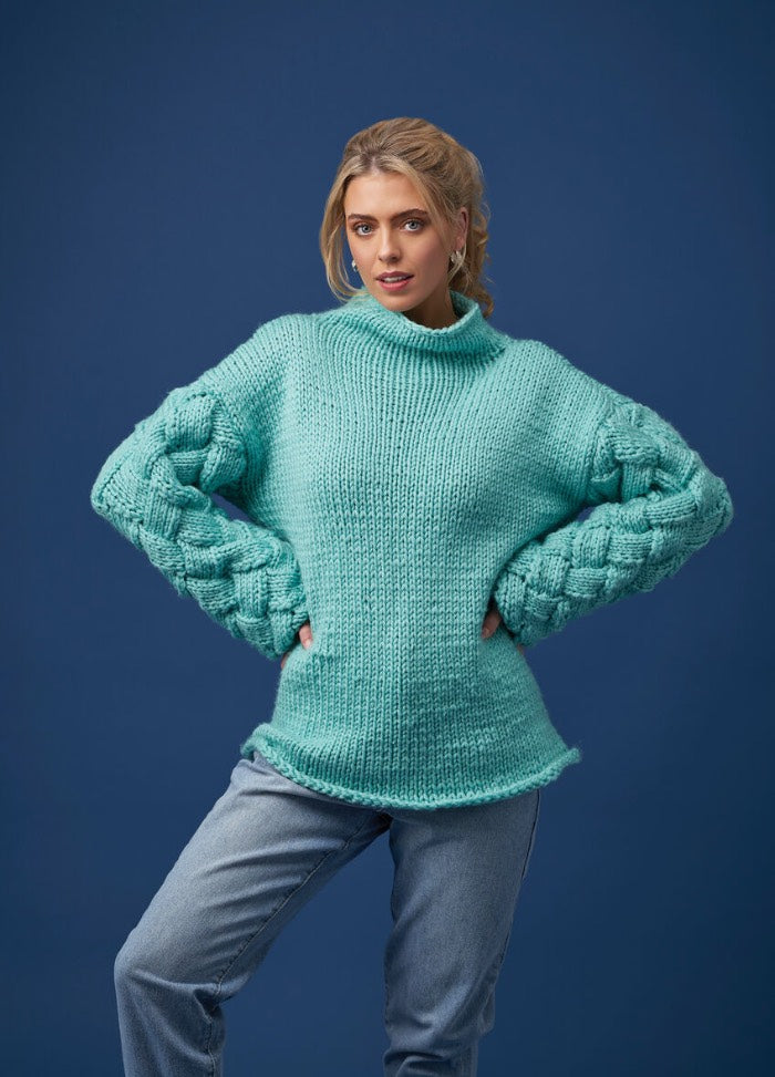 A woman poses confidently against a blue background in a textured turquoise sweater and jeans. Her hands rest on her hips, conveying a relaxed, self-assured vibe.