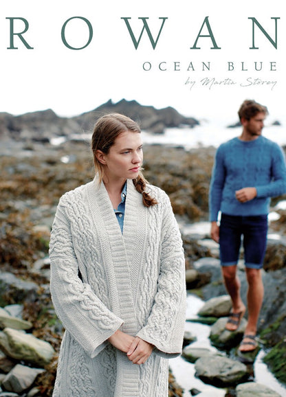Woman in a cream cable-knit cardigan stands on a rocky beach, looking thoughtful. A man in a blue sweater and shorts is slightly blurred in the background.