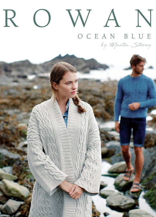 Woman in a cream cable-knit cardigan stands on a rocky beach, looking thoughtful. A man in a blue sweater and shorts is slightly blurred in the background.