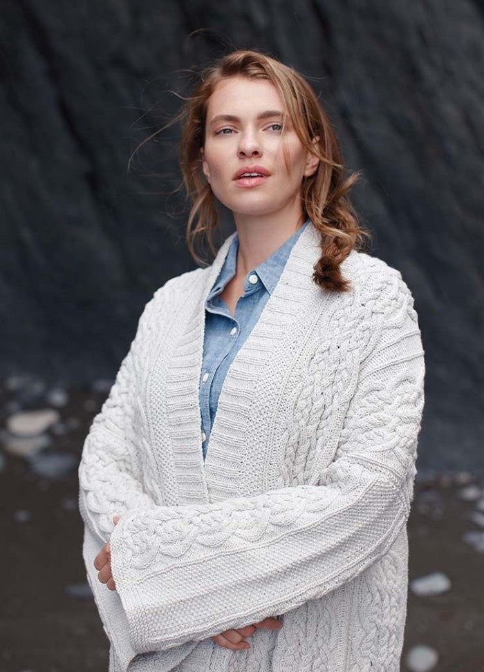 Woman with light brown hair and a calm expression stands against a dark rocky background. She is wearing a white cable-knit cardigan and a blue shirt.