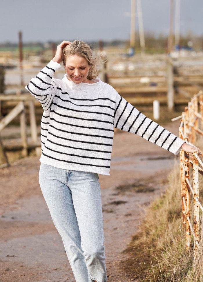 A person in a striped sweater and jeans walks on a rustic path, smiling with a hand in their hair. The scene is relaxed, with farm fencing in the background.