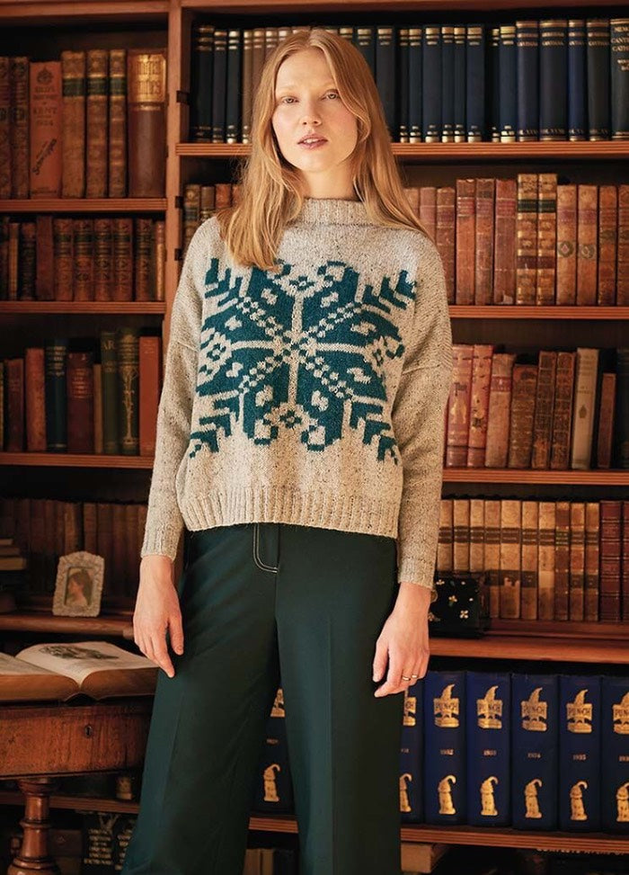 A woman in a cozy gray sweater with a large blue snowflake pattern stands in a library. She is surrounded by shelves of antique books, creating a warm and intellectual atmosphere.