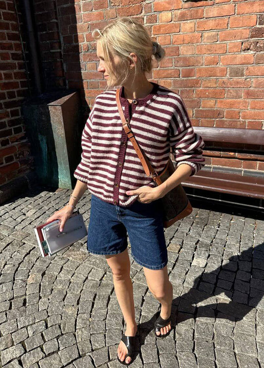 Person wearing the Olga Jacket with maroon and white horizontal stripes, styled with denim shorts, black sandals, and a brown crossbody bag, standing on a sunlit cobblestone street.