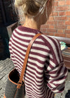 Back view of the Olga Jacket in maroon and white horizontal stripes, worn with a brown leather strap bag, outdoors against a brick wall.