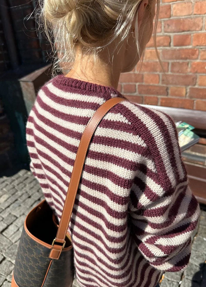 Back view of the Olga Jacket in maroon and white horizontal stripes, worn with a brown leather strap bag, outdoors against a brick wall.