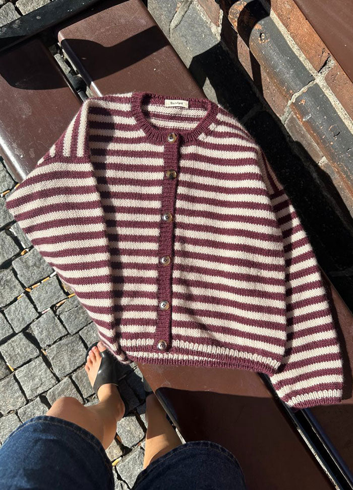 Flat lay of the Olga Jacket in maroon and white horizontal stripes with button-down front and ribbed edges, displayed on a bench with cobblestone background.