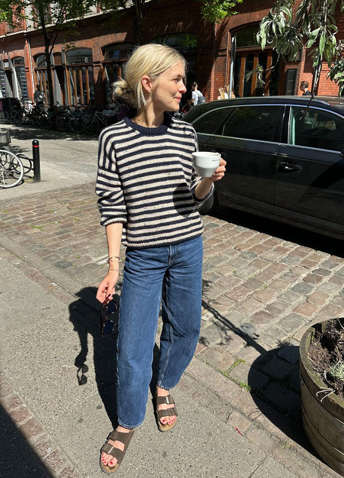 Person wearing the Olga Sweater in black and white stripes, styled with blue jeans and brown sandals, standing on a cobblestone street with bicycles and brick buildings in the background.
