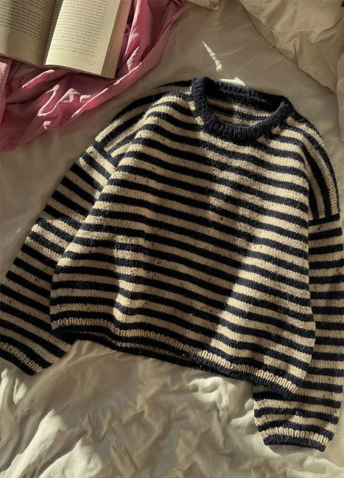 Flat lay of the Olga Sweater in black and white stripes with round neckline and long sleeves, displayed on a bed with white sheets and a pink blanket.