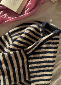 Flat lay of the Olga Sweater in black and white stripes, displayed on a sunlit bed with an open book and pink fabric in the background.