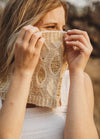 A woman playfully hides her face with a cream knitted scarf adorned with gold beads. Her eyes are visible, and she stands in warm, soft sunlight outdoors.