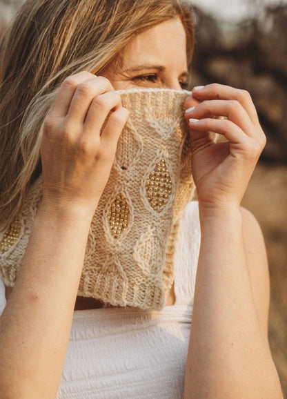 A woman playfully hides her face with a cream knitted scarf adorned with gold beads. Her eyes are visible, and she stands in warm, soft sunlight outdoors.