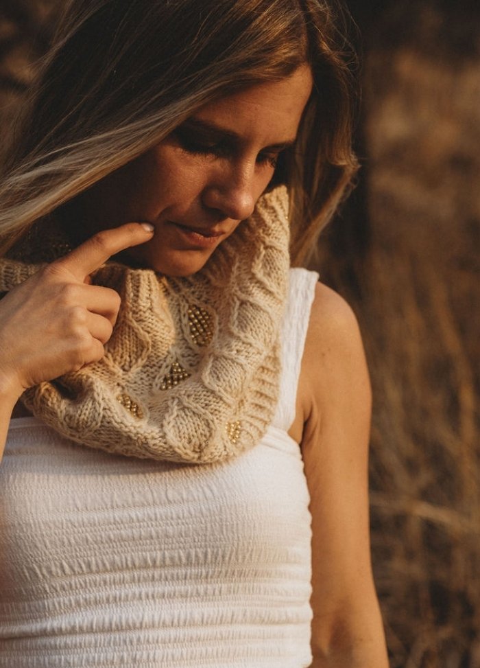 A woman in a white sleeveless top and knit scarf with beaded details stands thoughtfully outdoors. Warm sunlight enhances the contemplative mood.