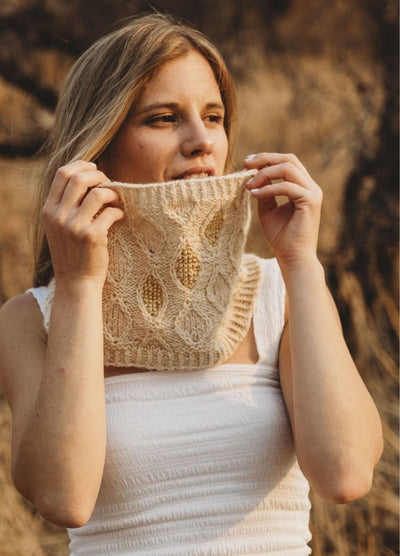 A woman in a white sleeveless top holds a beige knitted cowl with intricate patterns and gold accents, set against a blurred outdoor, sunlit background.