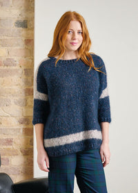 Woman with long red hair in a cozy blue sweater with white stripes, wearing plaid pants, stands in front of a brick wall, smiling softly.