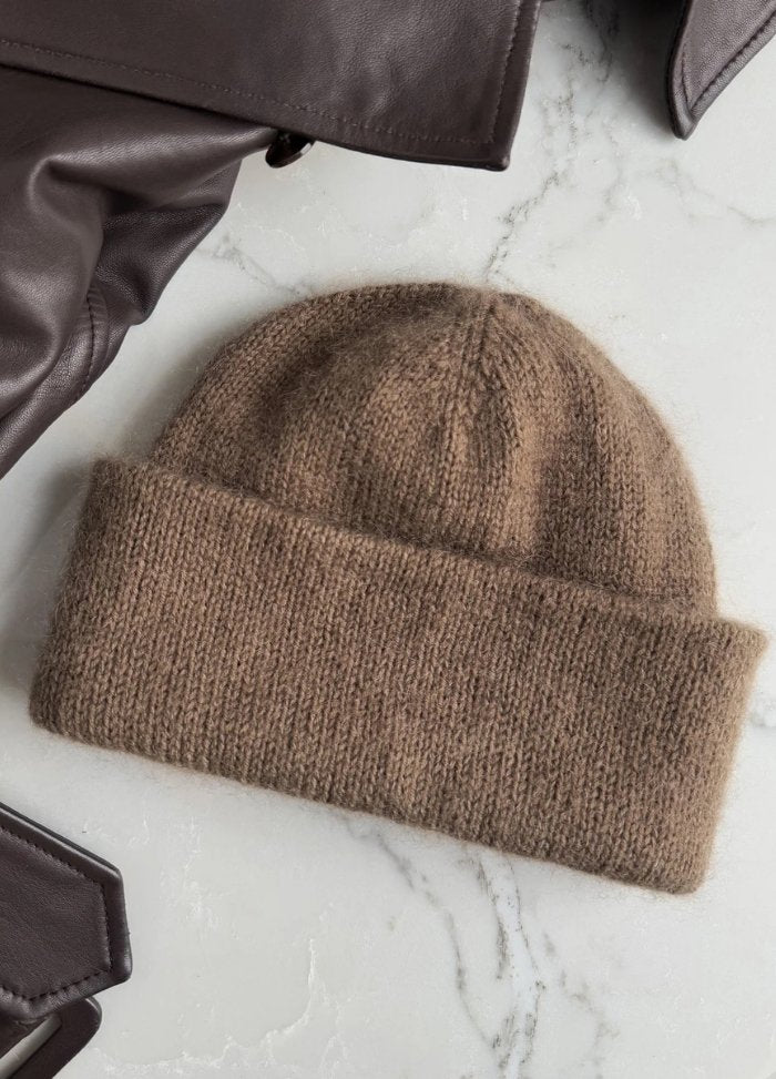 A cozy brown knit beanie lies on a marble surface next to folded brown leather jackets. The scene conveys warmth and style for cold weather.