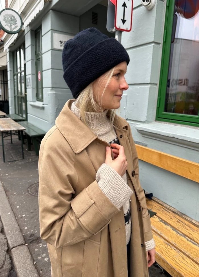 A woman in a tan coat and black beanie stands on a city street. She looks to the right, creating a thoughtful mood. There's a green window and a bench behind her.