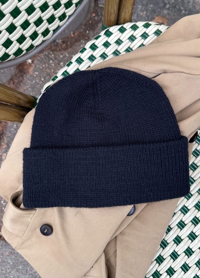 A navy blue knit beanie is placed on a tan coat, which is draped over a green and white woven chair. The setting has an outdoor, casual vibe.