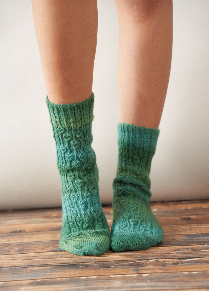 Close-up of a person's legs wearing hand-knitted green patterned socks standing on wooden floorboards. The scene conveys warmth and coziness.