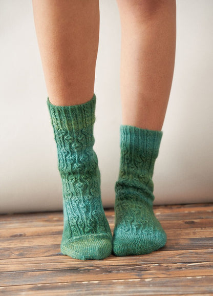 Close-up of a person's legs wearing hand-knitted green patterned socks standing on wooden floorboards. The scene conveys warmth and coziness.