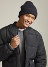 Smiling person in a winter outfit. They wear a black knit beanie, a dark quilted jacket, and a gray sweater, exuding a cheerful and warm vibe.