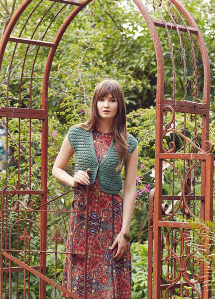 Model wearing the Pema Vest from the Rowan Softyak DK Collection by Marie Wallin, a green sleeveless knitted vest layered over a geometric patterned dress, photographed in a garden setting with a decorative rust-colored metal archway and lush greenery.
