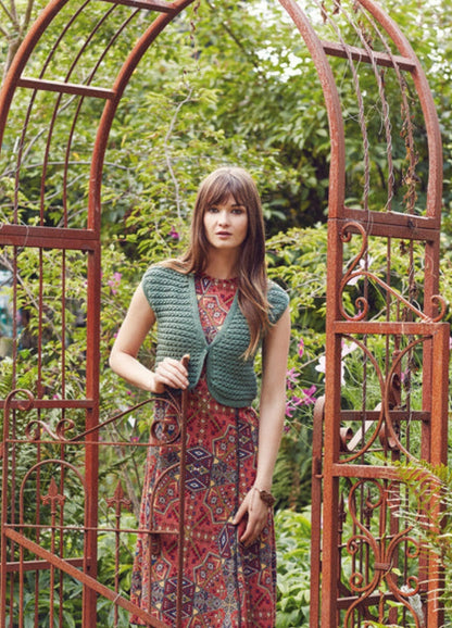 Model wearing the Pema Vest from the Rowan Softyak DK Collection by Marie Wallin, a green sleeveless knitted vest layered over a geometric patterned dress, photographed in a garden setting with a decorative rust-colored metal archway and lush greenery.