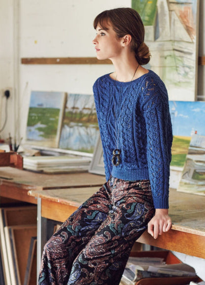 Model wearing the Posee Sweater from the Rowan Softyak DK Collection by Marie Wallin, a blue knitted sweater with intricate stitch patterns, paired with loose patterned pants, photographed in an art studio with landscape paintings and canvases in the background.
