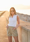 Woman stands against a sunlit stone wall wearing sunglasses, a white sleeveless top, and beige shorts. The setting sun casts a warm glow.