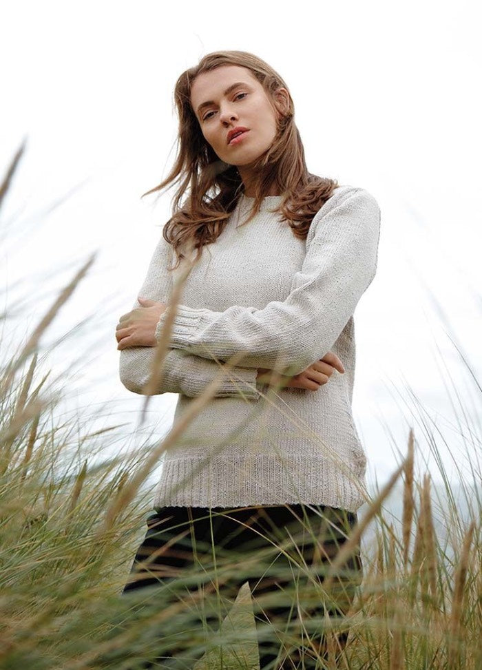 Young woman stands confidently in tall grass, wearing a light sweater. She looks thoughtful, with an overcast sky behind her. The mood is calm and introspective.