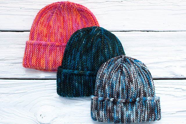 Paper Bag Caps displayed on a white wooden surface: one in pink and orange, one in dark green, and one in blue, gray, and white.