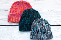 Paper Bag Caps displayed on a white wooden surface: one in pink and orange, one in dark green, and one in blue, gray, and white.