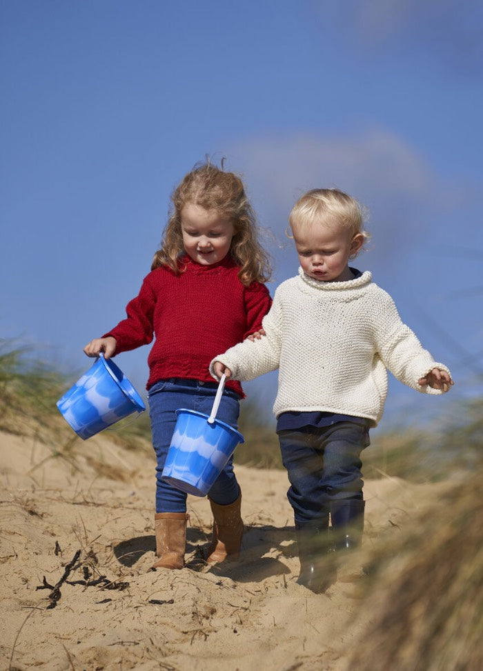 Two young children walk on a sandy beach under a clear blue sky, each holding a blue bucket. One wears a red sweater, the other a white one, evoking joy and exploration.
