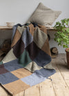 Cozy scene with a patchwork blanket in earthy tones draped over a bench, accompanied by a beige cushion. A cup and potted plant add warmth.