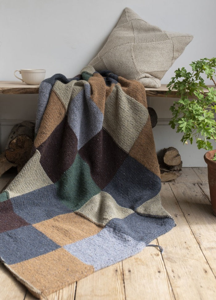 Cozy scene with a patchwork blanket in earthy tones draped over a bench, accompanied by a beige cushion. A cup and potted plant add warmth.