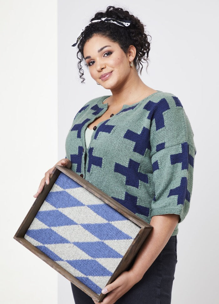 A person smiles while holding a wooden-framed geometric pattern artwork. They wear a green and blue sweater with crosses, exuding creativity and warmth.