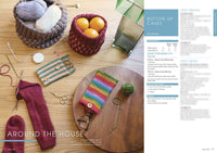 Wooden table with knitting supplies; a basket with yarn, another with oranges. Two knitted cases, red sock in progress, scissors, and needles. Cozy, crafty vibe.