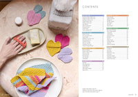 A flat lay of knitted accessories on a pale marble surface, including a hand holding an orange knitted soap holder with a bar of soap, a tissue box covered in a grey knitted cozy with small flower motifs, and heart-shaped knitted coasters in pink, yellow, and purple. A plate in the foreground holds folded pastel-striped knitted cloths. The right side of the image shows a contents page listing patterns under categories like Self Love Club, Around the House, Celebrate Baby’s Arrival, and more.